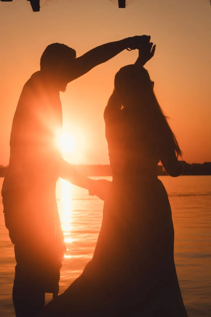 Silhouette of a couple dancing hand in hand by a lake at sunset, with the sun glowing between them.