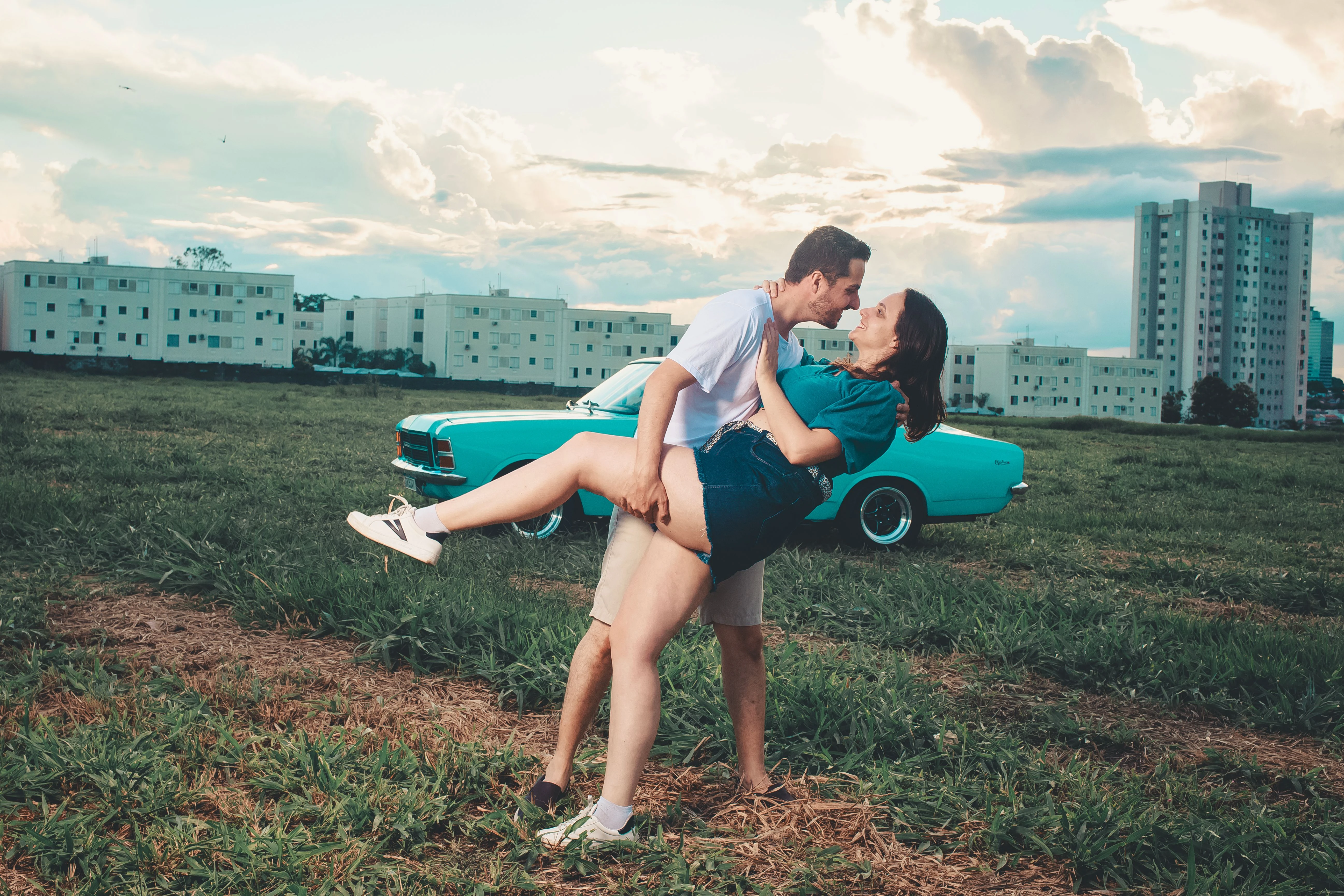 Man dipping his smiling partner in a playful dance pose in a grassy field, with a vintage teal car and apartment buildings in the background