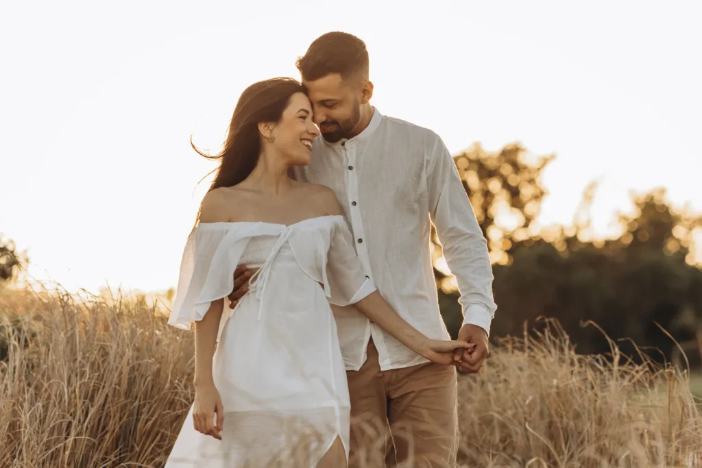 Smiling couple in white linen outfits embracing and holding hands in a golden field at sunset, sharing a tender moment of emotional intimacy.