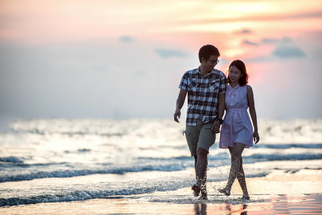 Couple holding hands and walking barefoot through shallow waves on a beach at sunset