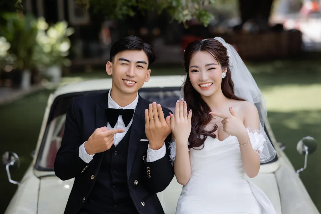 Newlywed bride and groom smiling and showing off their wedding rings beside a vintage car