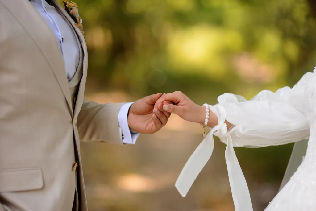 Groom in a beige suit gently holding the bride’s hand in a wedding outfit with a pearl bracelet